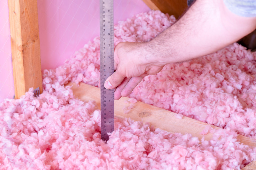 person measuring the depth of pink fiberglass blown insulation in an attic.